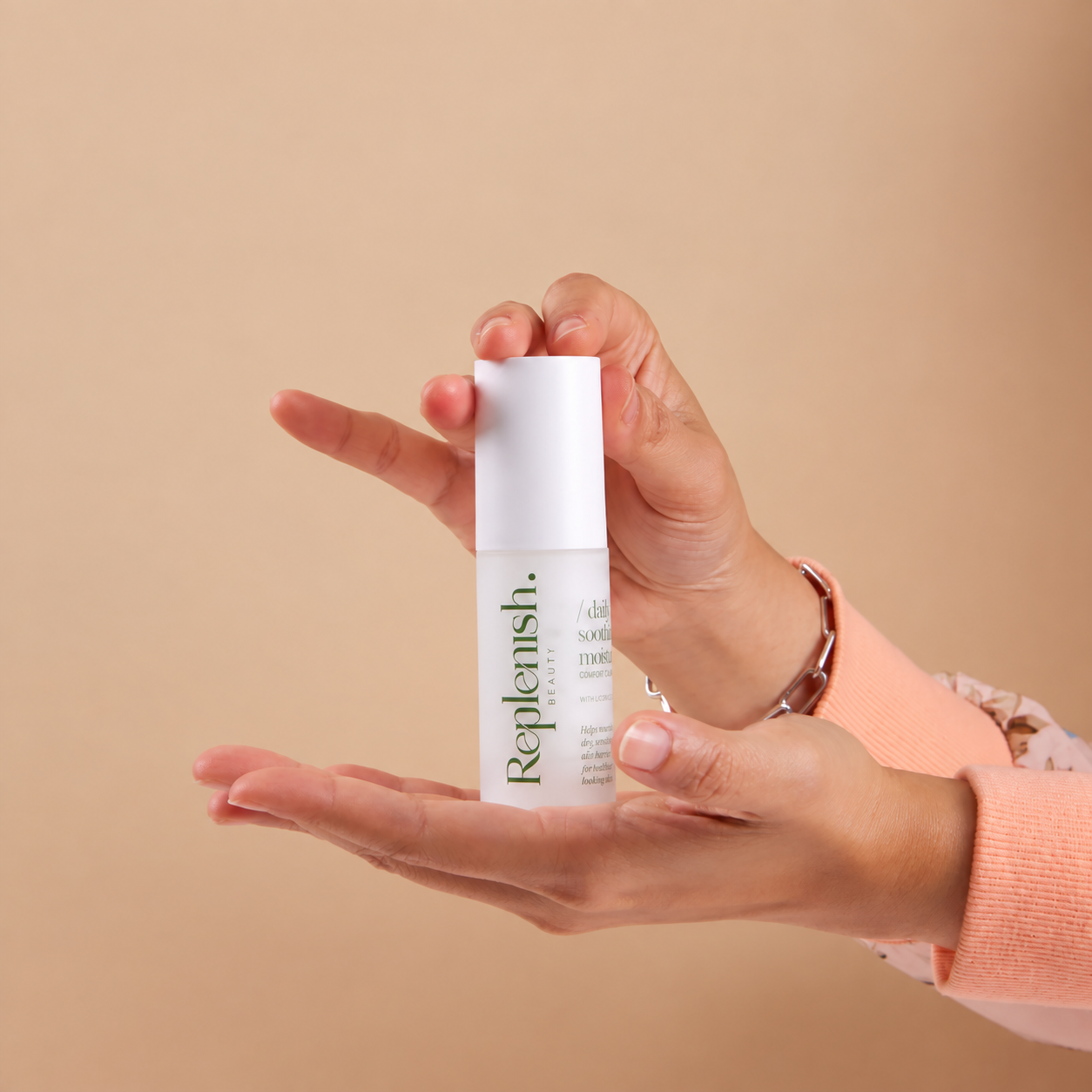 Two hands holding Replenish Daily Soothing Moisturizer against a light peach background